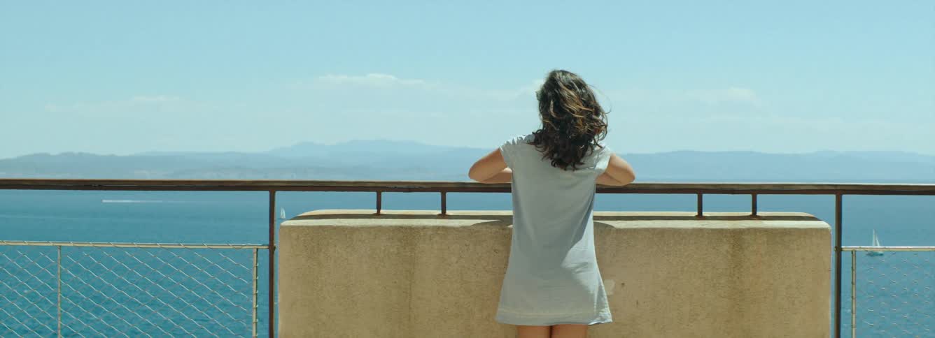 A woman walks up to lean against a railing and look at the sea in Bonifacio, Corsica