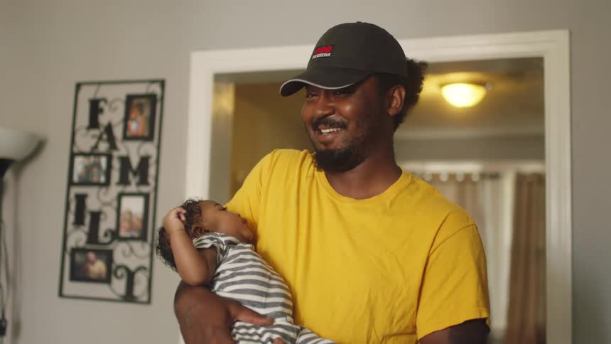 A man smiles into the camera while rocking a baby in his arms