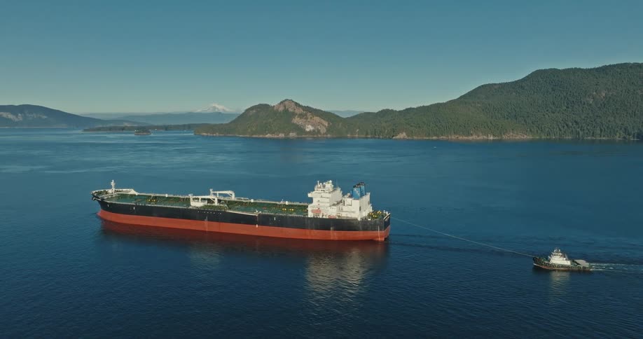 A large ship is sailing in the water and pulling a smaller boat behind it with mountains in the distance under a clear sky
