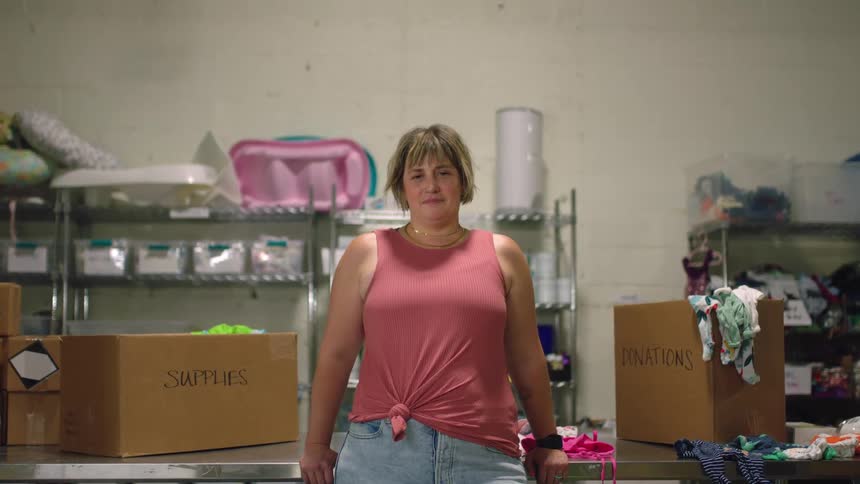 A woman is leaning against a table with boxes and shelves behind her