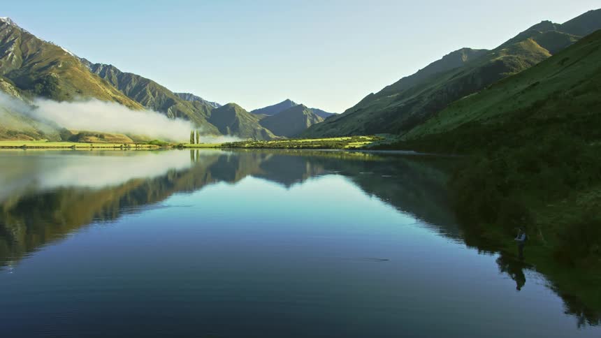 Fisherman stands on shore fishing as lake reflects sky and surrounding mountains in New Zealand