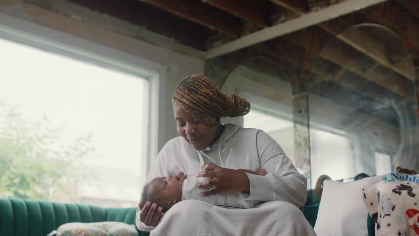 Mother with brown dreadlocks gray hoodie holds and kisses newborn baby in living room at home