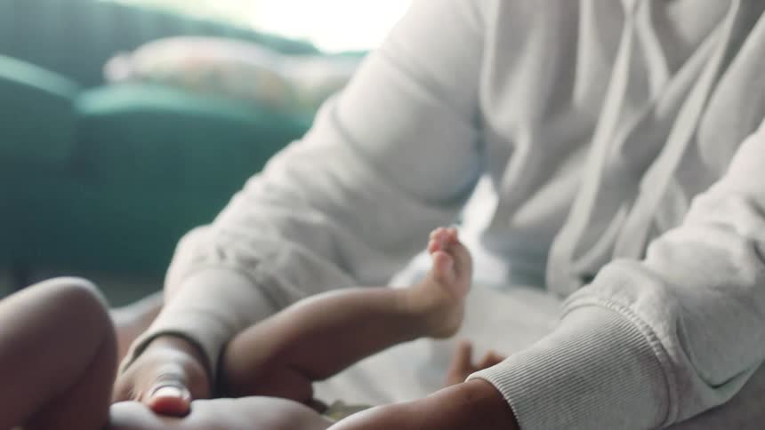 Woman with brown dreadlocks and gray hoodie rubs babys belly in living room at home