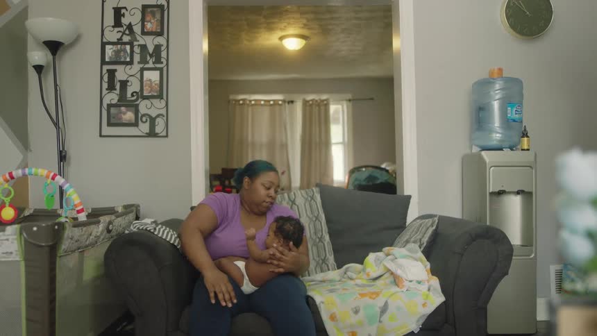 A woman sitting on a sofa in the living room of a house removes the shirt from a baby resting in her lap