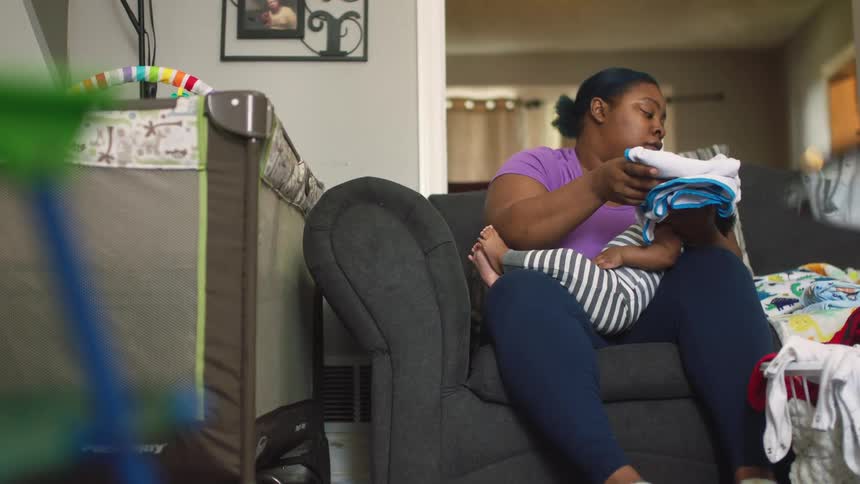 A woman sitting on a sofa in the living room of a house folds clothing while a baby sleeps in her lap