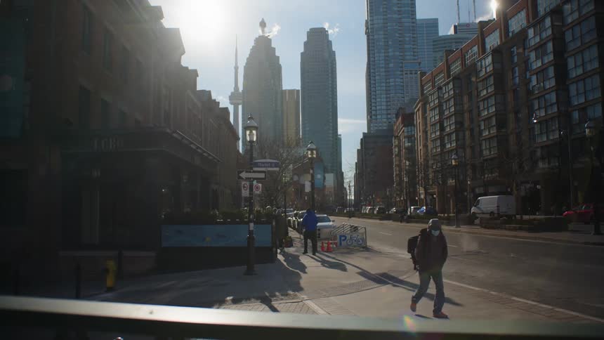 Pedestrian crosses intersection at crosswalk while others walk along sidewalk toward downtown skyscrapers on sunny day