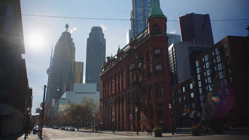 Pedestrians move along sidewalk near turreted building with city skyscrapers in the background on a bright sunny day