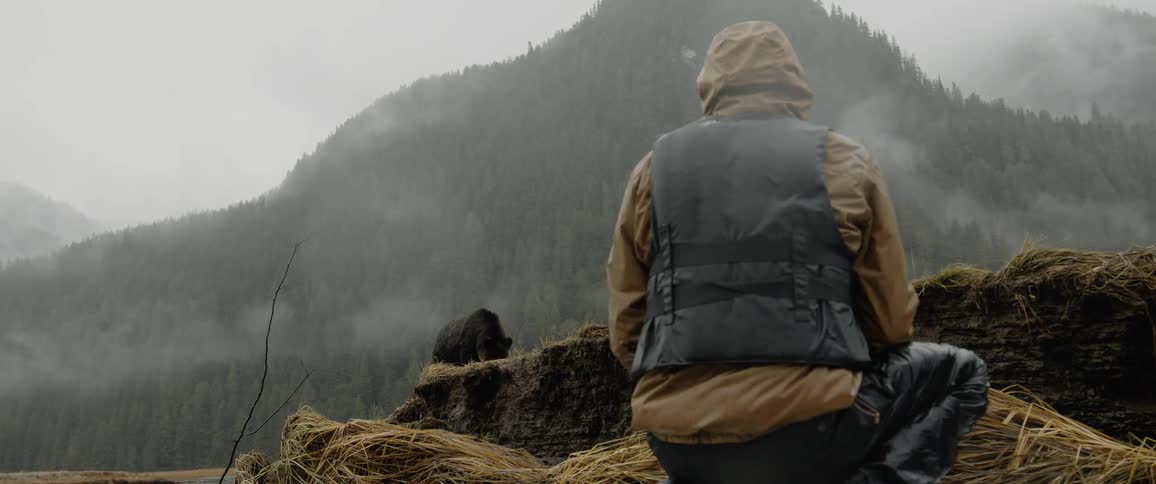 Photographer crouches taking picture of bear with mist covered forested mountains in the background