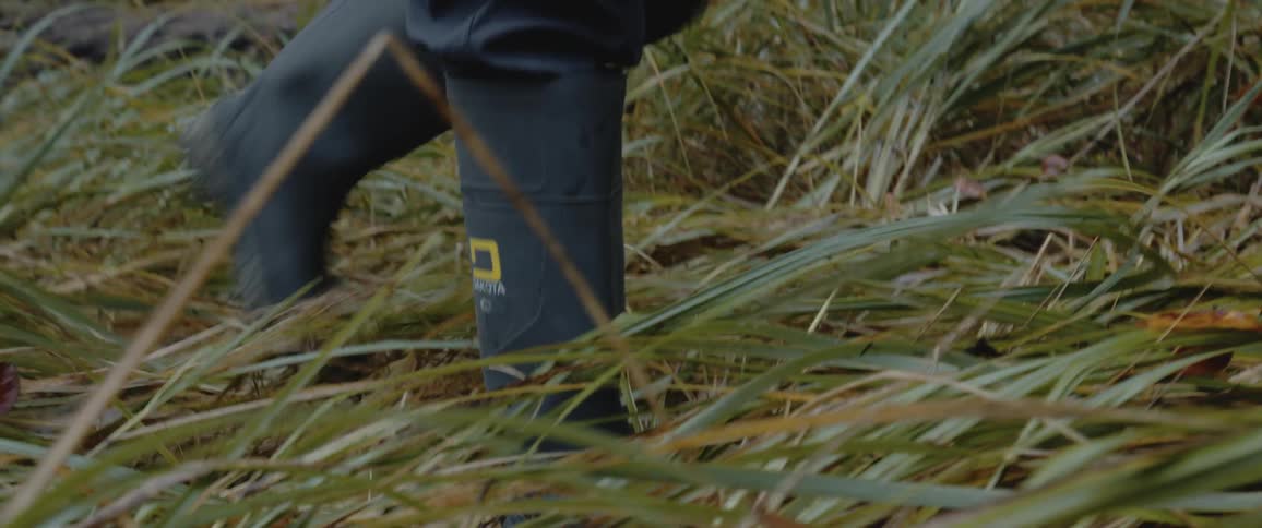 Person wearing pants tucked into boots walks across mostly flattened grass