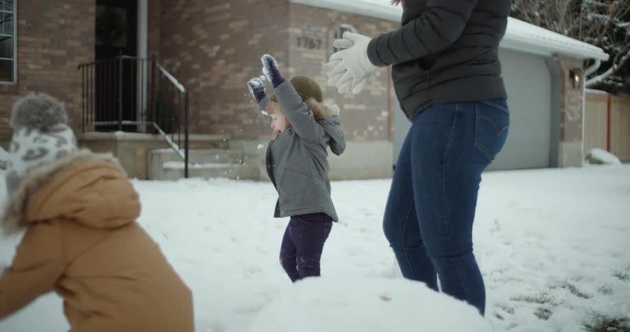 Two little girls throw snow outside their home while their mom watches