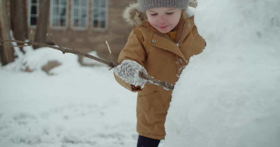 A girl sticks a branch into a snowman shes made in the front yard with her mom
