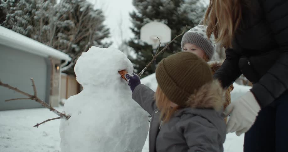 Mother helps her daughter to put a nose on a snowman they built in the backyard at winter