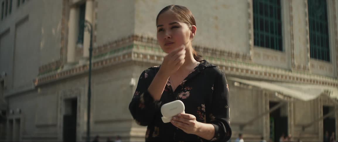 The sun shines on a woman standing on a city street as she opens a container and takes out earbuds and puts them in her ears