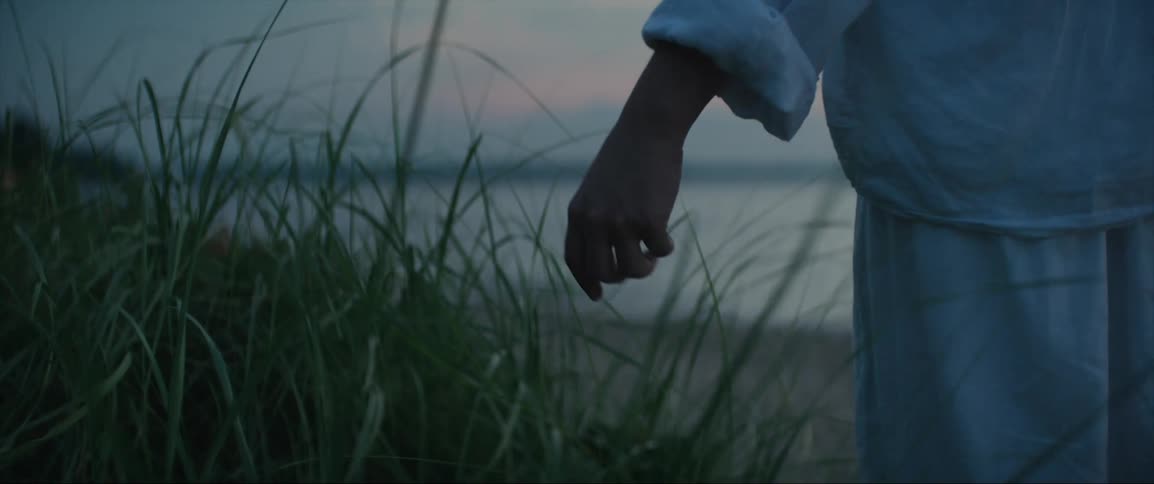 A woman is walking along the coast with her hand brushing over tall grass as she passes and waves roll into shore