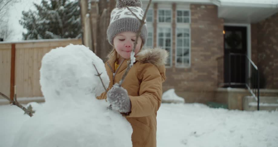 A young girl building a snowman in front of her house puts a branch in for the arm