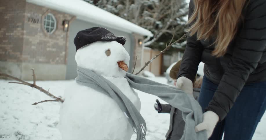 A woman outside her home puts a scarf on a snow man for her daughter