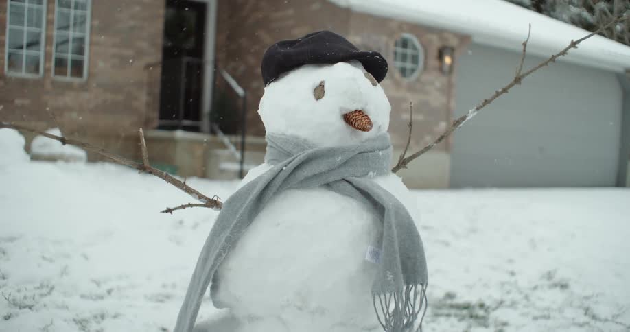 Snow falls on a snowman in front of a house