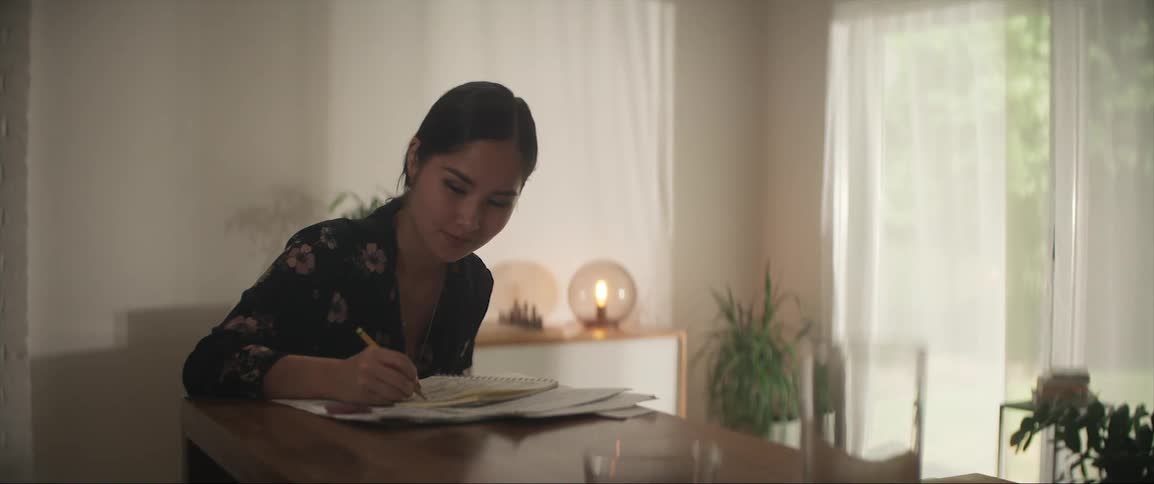 Woman with ponytail and blue shirt sits at table writing down in paper in the living room