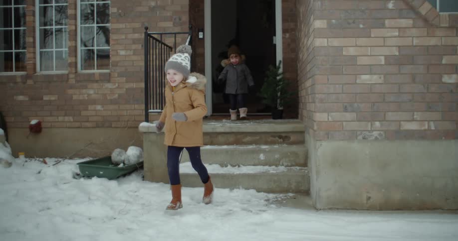 A young girl runs out of a house to play in the snow covering the front yard