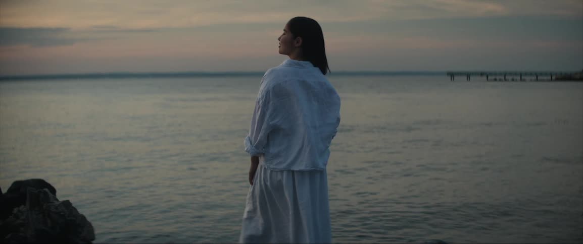 A woman is standing on rocks along the coast looking at the sky as waves roll into shore