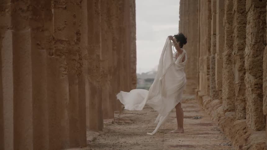 A woman holds out the edges of her dress to the wind while standing among ancient columns