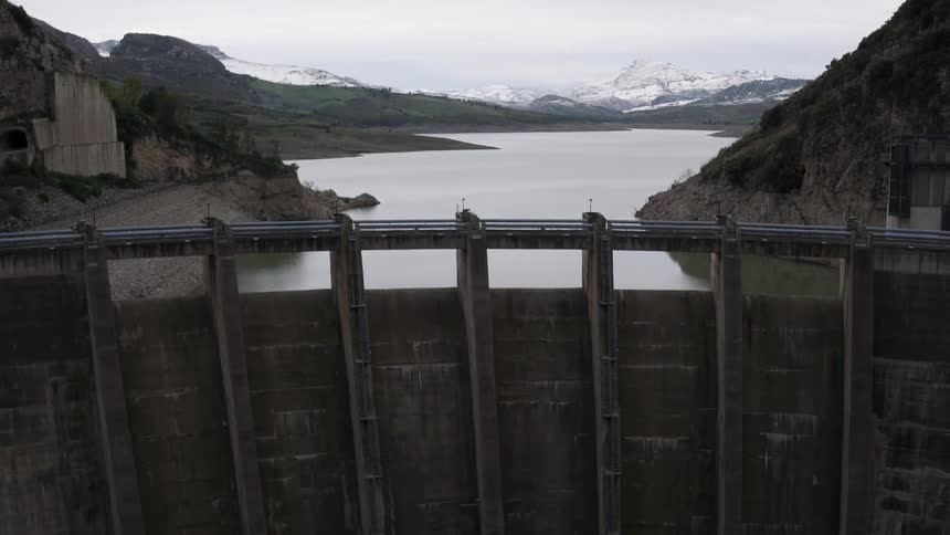 A large dam, a lake, and mountains are seen from an aerial view