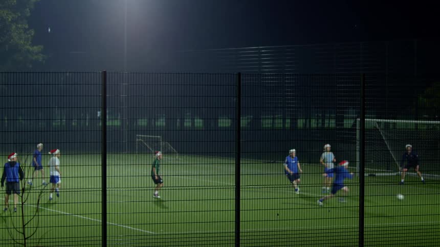Five a side soccer on a  floodlit pitch at night, seen through a wire mesh fence