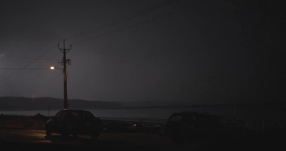 Automobiles are parked overlooking the coast as lightning flashes across the dark sky