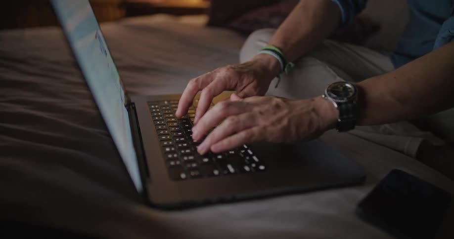A man types on his laptop while sitting in bed