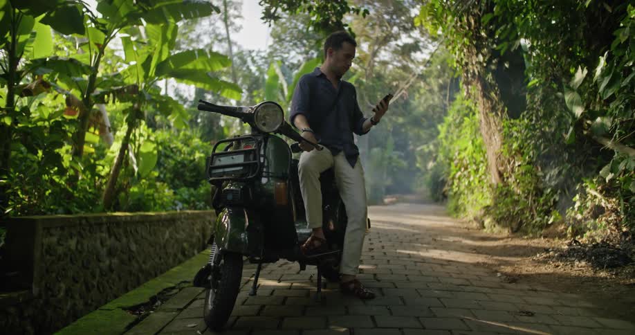 Man sitting on a motorcycle parked on a road on the tropics checking his smartphone