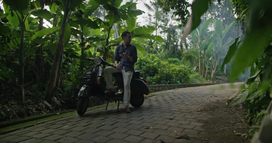 A man sitting on a parked moped looking at his phone and at the palm trees while waiting