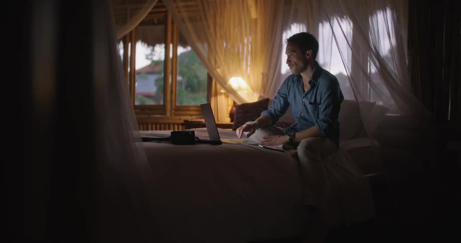 Man with blue shirt and brown pants sits on bed working on tablet in bedroom