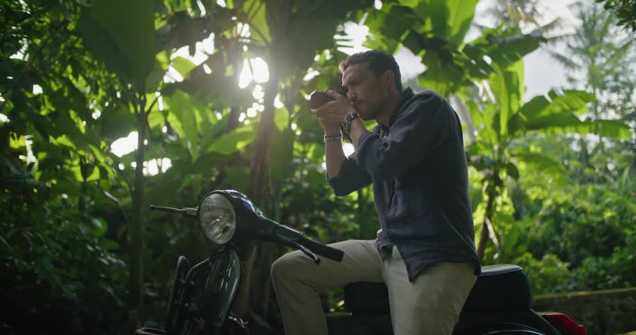 A photographer sitting on a moped parked on a bridge in a woodland raises his camera to take a photograph of his surroundings