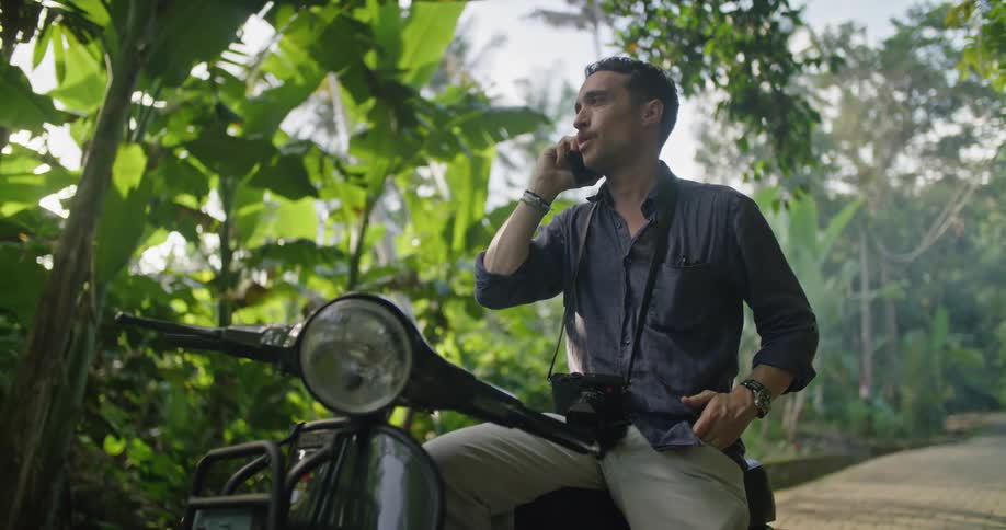 Man sitting on a motorcycle parked on a road on the tropics talking on his smartphone