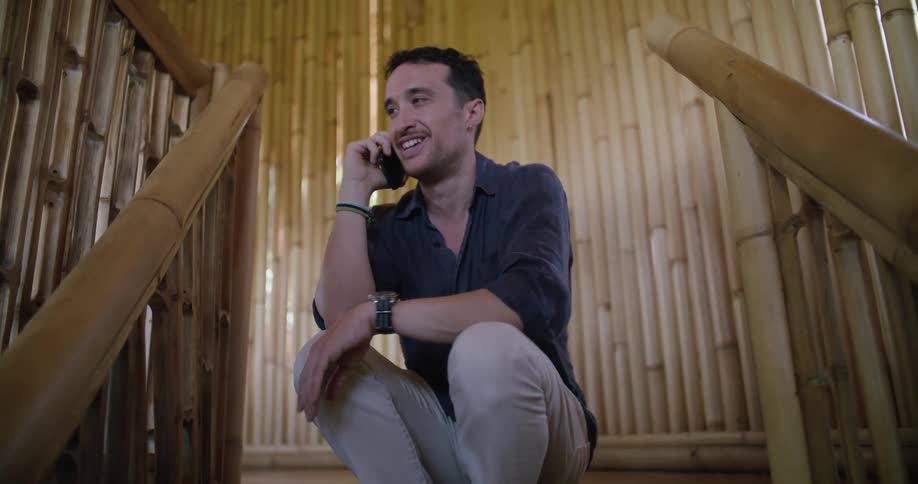 A man sits on the stairs and talks on the phone in a room built completely of bamboo logs
