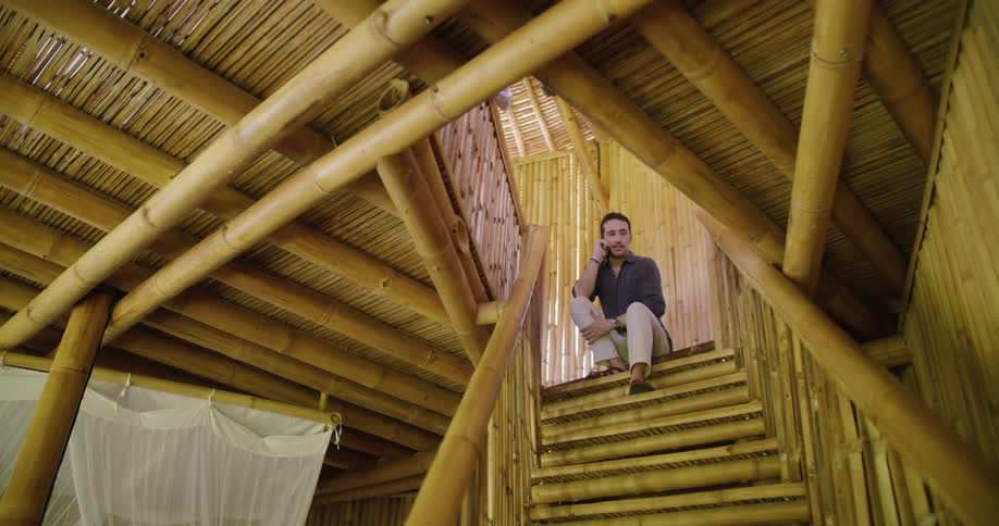 Man sits at top of staircase in building made of bamboo and makes a call