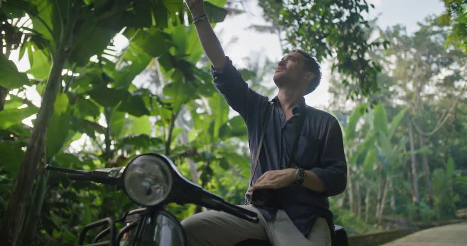 A man is sitting on his motorbike surrounded by tropical plants holding his phone and he raises it in the air and shakes his head