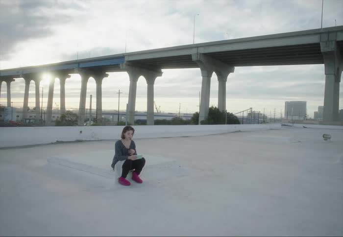 A woman sitting on a rooftop holding her phone and moving her arm and hand to music
