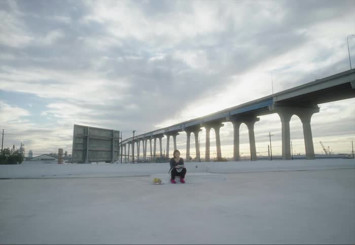 Woman crouches reading near elevated highway as red tinted sky and city skyscrapers sit on the horizon