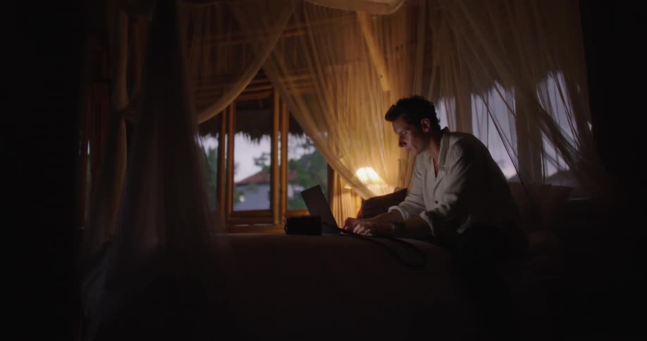 Man with blue shirt and brown pants sits on bed working on laptop and camera in dim lit room