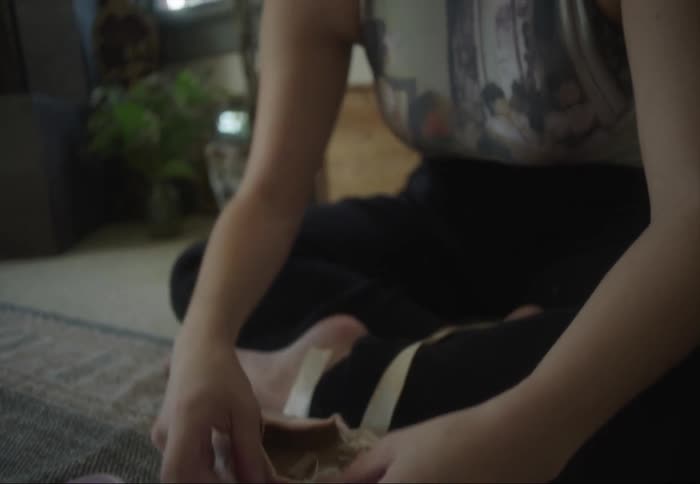 Young woman sitting on the living room floor sewing a pair of ballet toe shoes