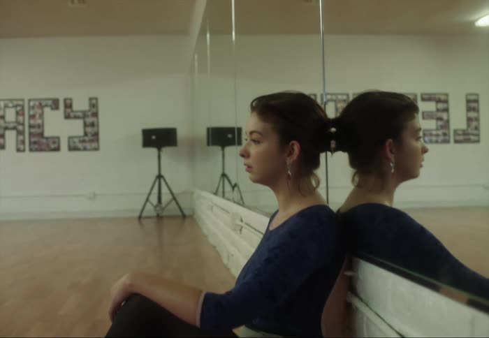 Ballerina sits on floor of dance studio leaning against wall covered by mirrors