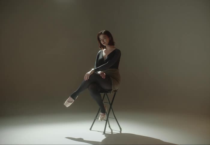 A woman is sitting on a stool with her legs crossed as cameras flash and she moves her hair back with her hand