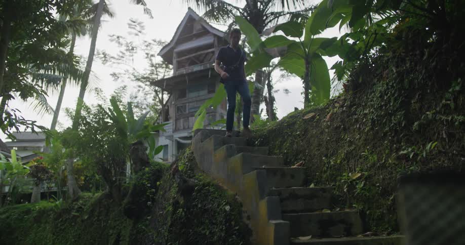 Young man carrying a camera walks down the stairs on the side of a hill on a tropical rural area
