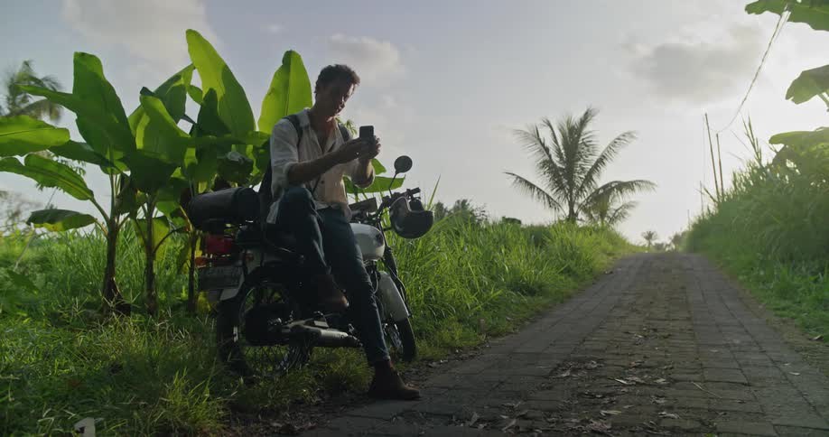 A man is sitting on his motorbike on the side of a cobblestone road holding his cell phone and looking around