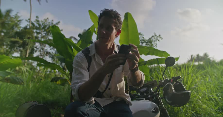 A man is sitting on his motorbike holding his cell phone with tropical plants behind him