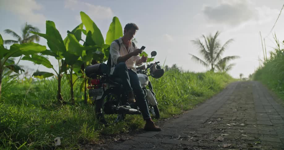 A man is sitting on his motorbike on a cobblestone road using his cell phone and looking around