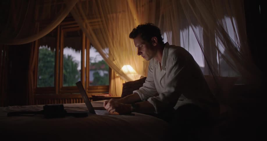 A man is sitting down typing on a laptop computer as a lamp shines near open windows
