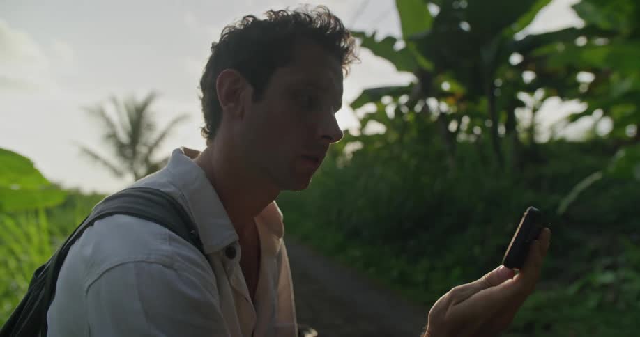 A man is standing on the side of a dirt road surrounded by tropical plants and using his cell phone