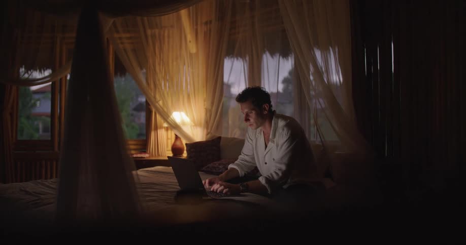 A man sits down a camera and begins typing on a laptop while sitting on a bed in a dimly lit bedroom